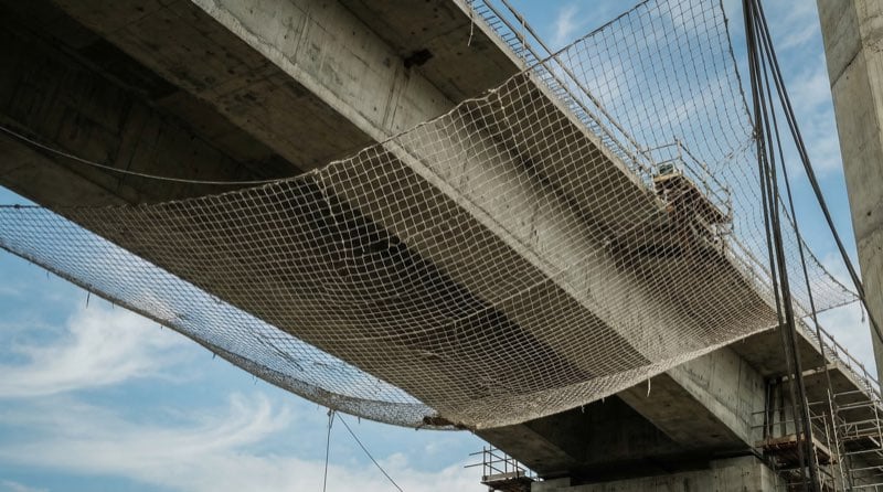 Wide shot looking upward at a construction safety net stretched beneath a structure, showing the net against concrete and sky