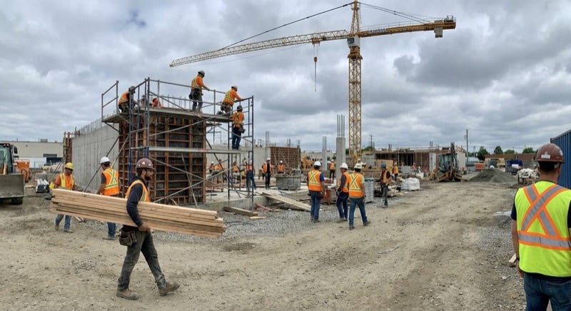Busy commercial construction site with crane, scaffolding, and multiple trades workers in hi-vis vests and hard hats