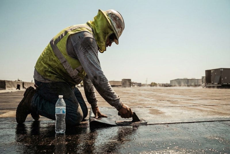 Roofer working on commercial flat roof in intense summer heat with heat shimmer rising from dark roof surface