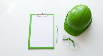 Safety meeting preparation materials including a green clipboard, hard hat, safety glasses, and pen on a clean white desk
